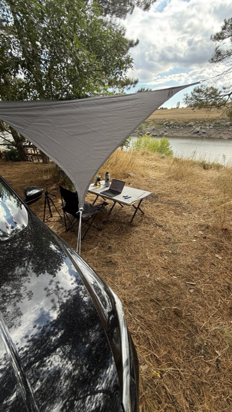 A camping table with a laptop by a river, car and tarp behind it.